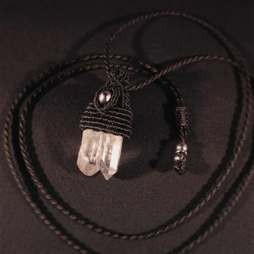 Ett svart halsband i makramé med en dubbel bergkristallspets och pärlor i rostfritt stål, vilandes mot en mörk bakgrund.