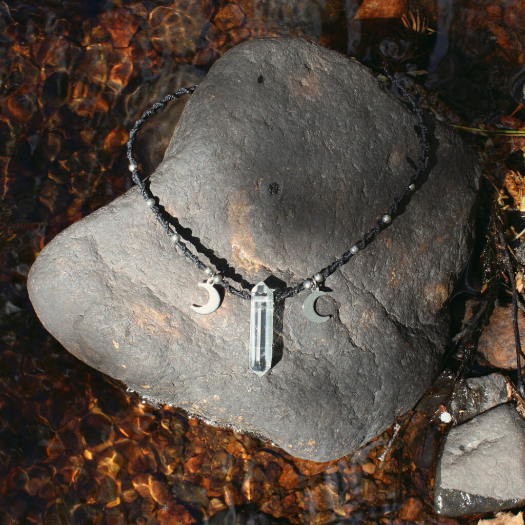 Ett svart halsband med bergkristallspets och månar. Halsbandet är knutet med makramé i svart tråd och har också metallpärlor i rostfritt stål.