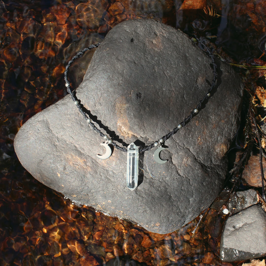 Ett svart halsband med bergkristallspets och m&aring;nar. Halsbandet &auml;r knutet med makram&eacute; i svart tr&aring;d och har ocks&aring; metallp&auml;rlor i rostfritt st&aring;l.