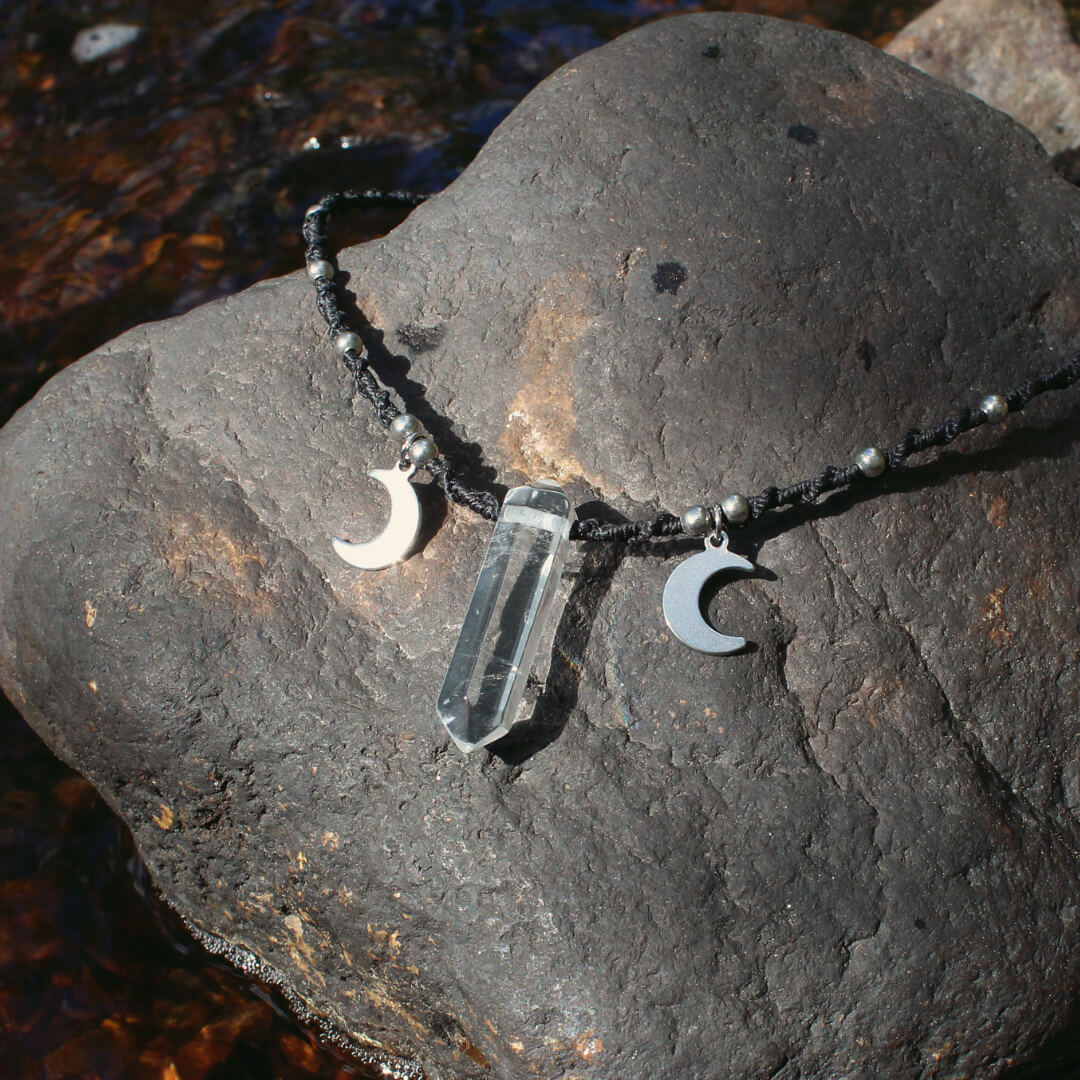 N&auml;rbild av ett svart halsband med ett h&auml;nge av bergkristall och m&aring;nar p&aring; sidorna om kristallen. Halsbandet ligger p&aring; en sten.